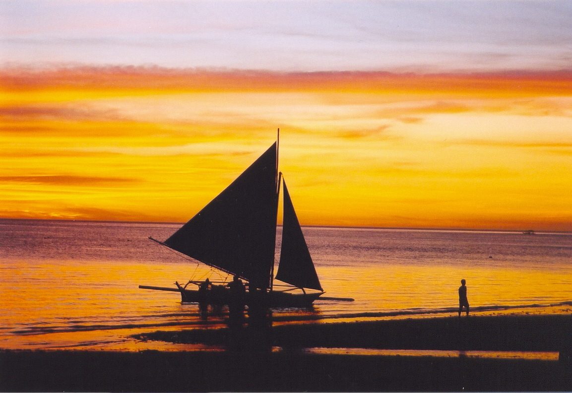 Traditional paraw outrigger sailboat with white sails off Boracay's White Beach at sunset