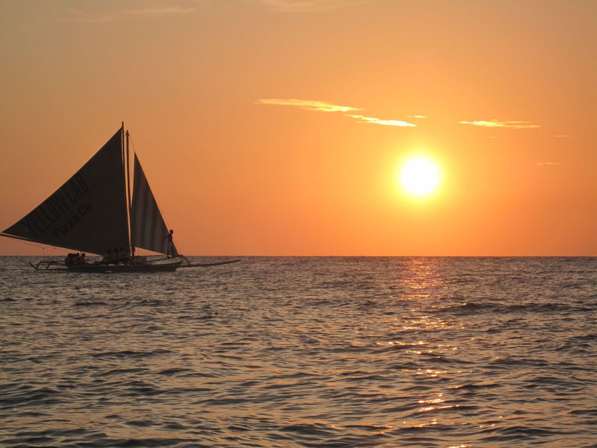 A traditional paraw sailboat with outriggers silhouetted against the Boracay sunset off White Beach